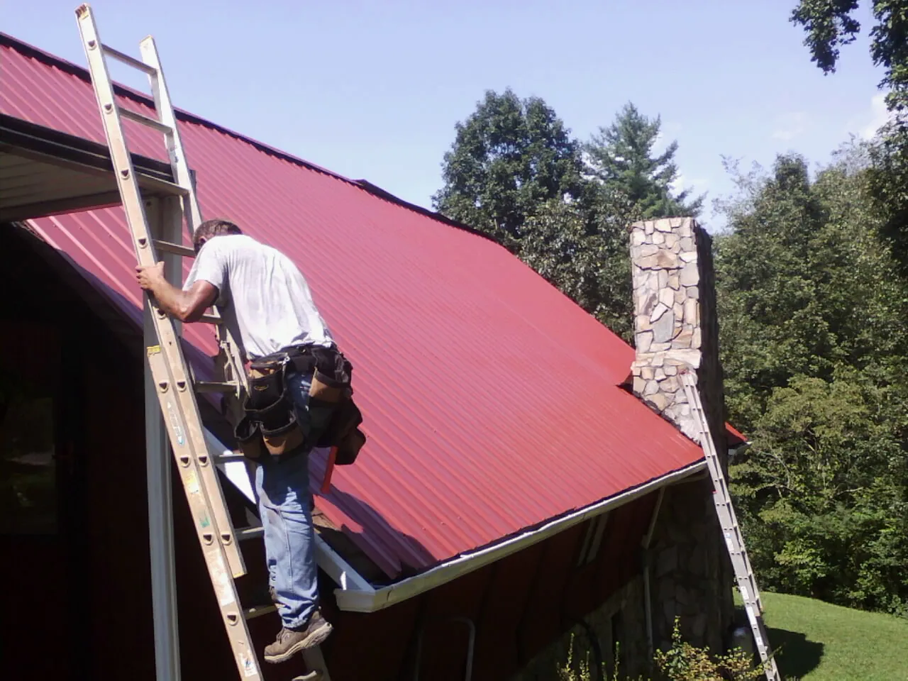 Roofer on a ladder beside a red metal roof and stone chimney — Soffit & Fascia Repair by Roofing Brothers in Greenfield Center